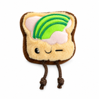 Juguete de peluche para perros marca Verný con forma de tostada animada. Tiene una cara sonriente que guiña un ojo, detalles de cobertura de mantequilla y rebanadas de aguacate en la parte superior, y dos patas colgantes de cuerda marrón